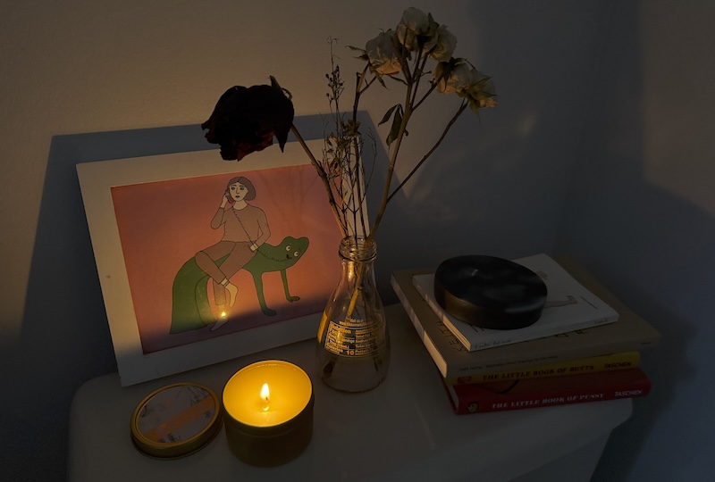 photo of a pile of books, framed art, vase with dried flowers, and a lit tin candle