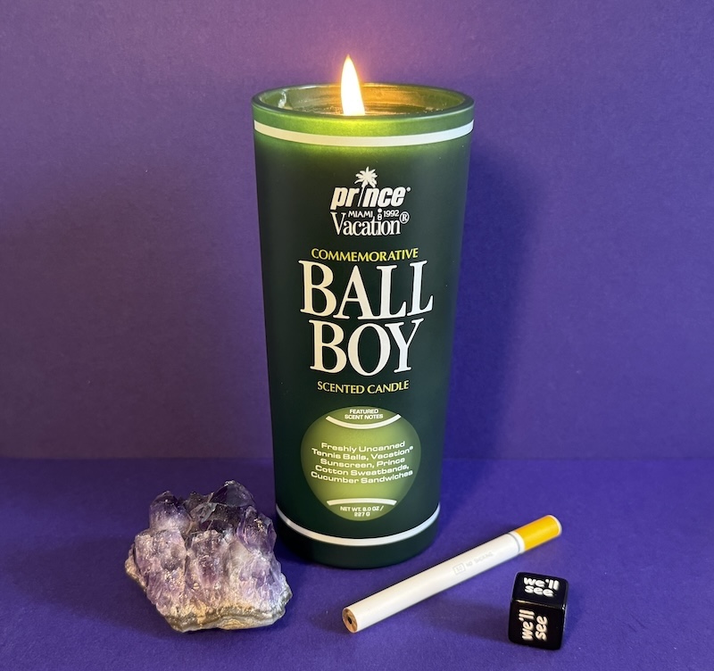 photo of the prince vacation ball boy scented candle that's in a 8 oz green tumblr glass. alongside of it on and in front  of a purple backdrop are an amethyst crystal, cigarette pencil and a die where each face says "we'll see"