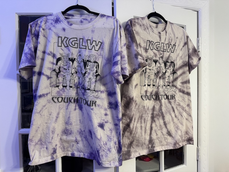 two of our kglw couch tour band shirts hanging up on a door. they are both tie dyed, one in hyacinth purple and the other in charcoal gray