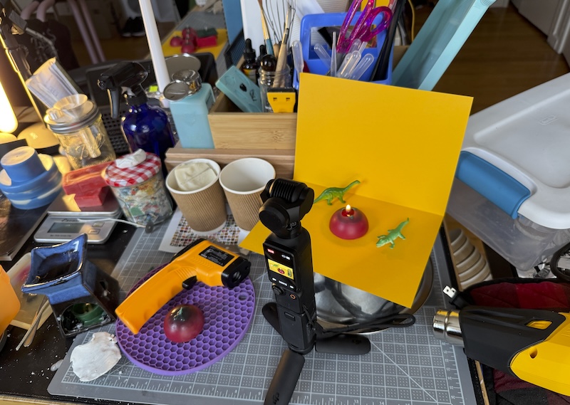a photo of my messy candlemaking area and i'm recording a burn test of a tomato sample candle on a goldenrod paper backdrop with a couple little figurines. it's an aesthetic bubble amidst the chaos of the rest of my desk