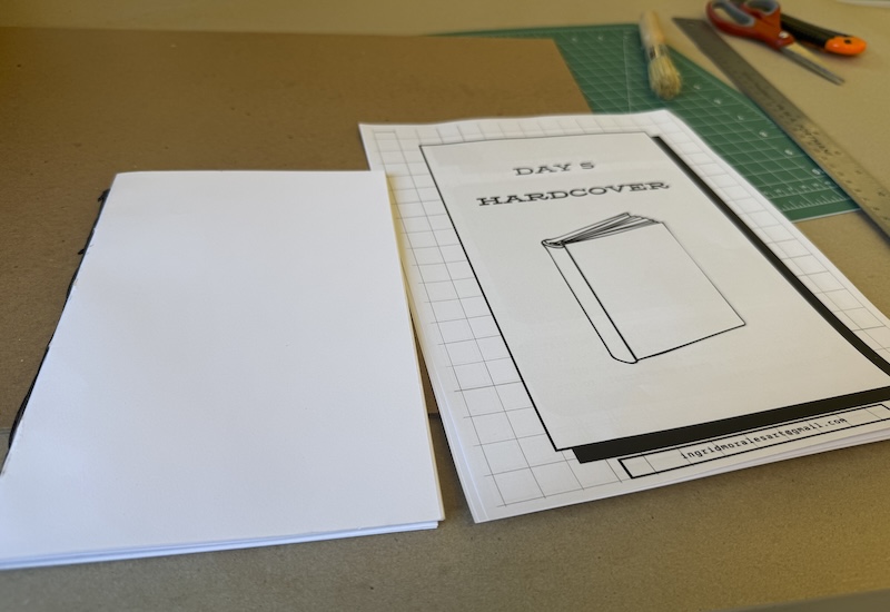 photo of a table at the bookbinding workshop with a "day 5 hardcover" packet, hard board, a cutting mat, ruler, glue brush, my textblock of paper, scissors and a box cutter