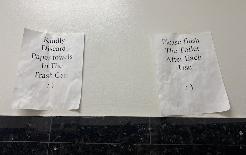 photo of two printer pages on the bathroom wall. one says "Kindly Discard Paper towels In The Trash Can : )" and the other says "Please flush The Toilet After Each Use : )"