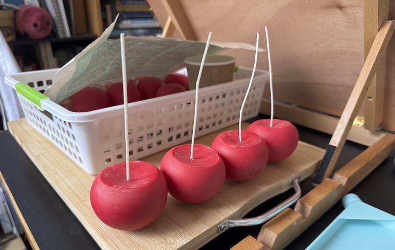 photo of one end of my candle making area of my desk. there are four wicked, red sphere candles and my bookshelf is in the background