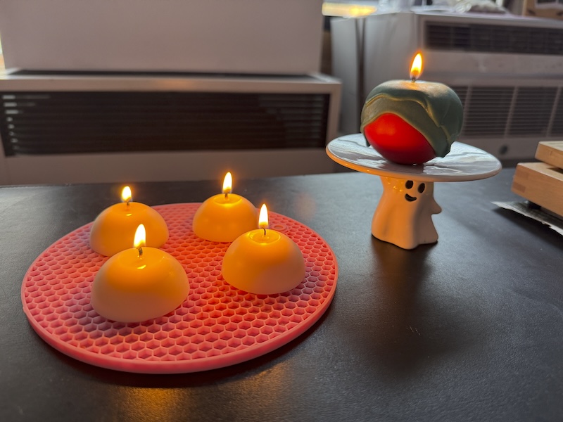 photo of a wick test of four white small half spheres and on a ghost dish (a cute ghost holding up the dish part)  there’s an apple shaped candle with green ooze on it lit