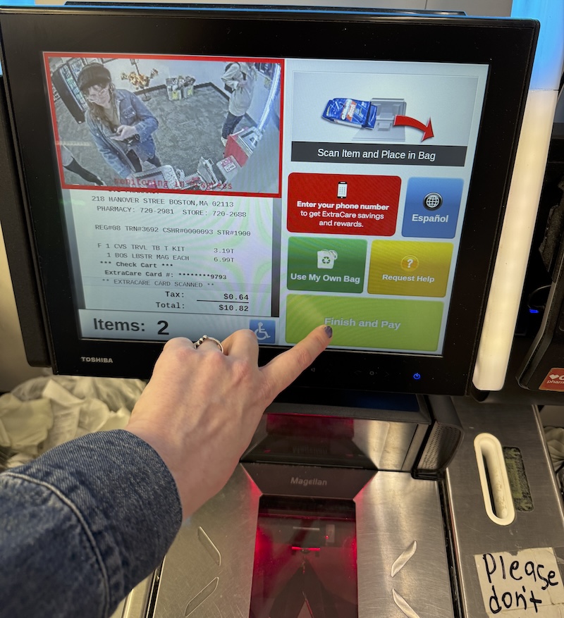 photo of my hand checking myself out on a self-checkout line in cvs, it shows surveillance of me in front of the register. i'm buying toothpaste and a lobster souvenir magnet
