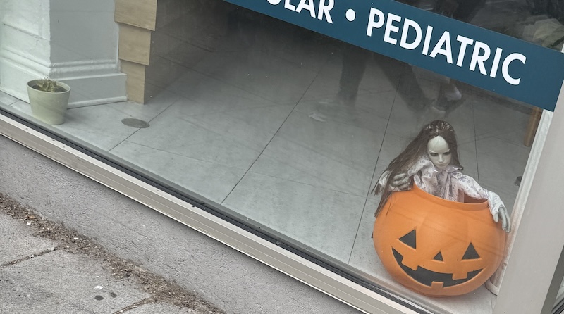 photo of the bottom of a window for a physical therapy office on the street that, below a window banner that says "pediatric" among other services is a scary doll and jack o'lantern