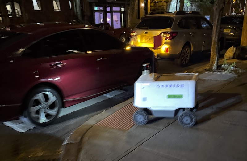 blurry night photo of an uber eats robot trying to cross at the corner but there's a car parked on the crosswalk with its hazards on