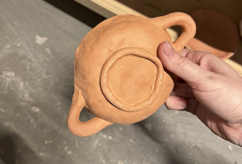 my dusty hands holding up a single-person-sized casserole dish made of red clay that i have yet to glaze