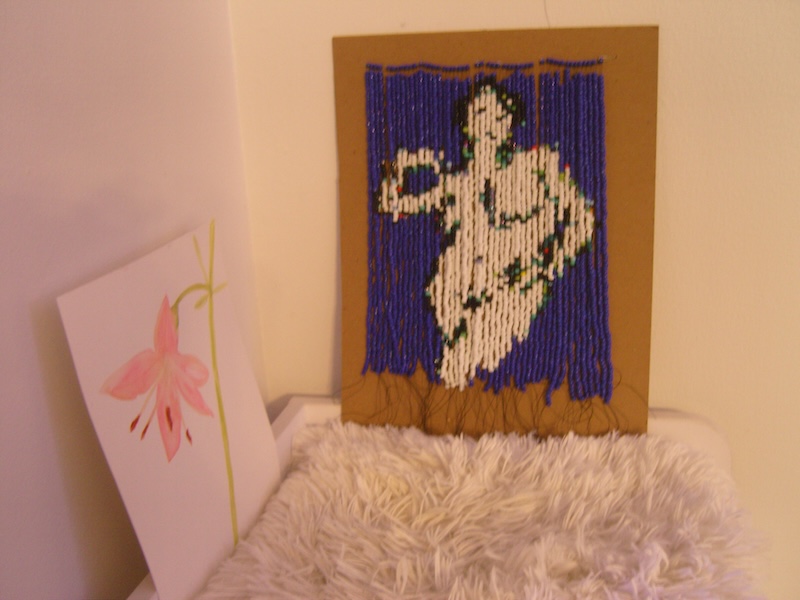 photo of a printer-paper-sized beaded tapestry where the strands are mounted from the top to a thick piece of brown board and the image is the same figure and colors as the painting