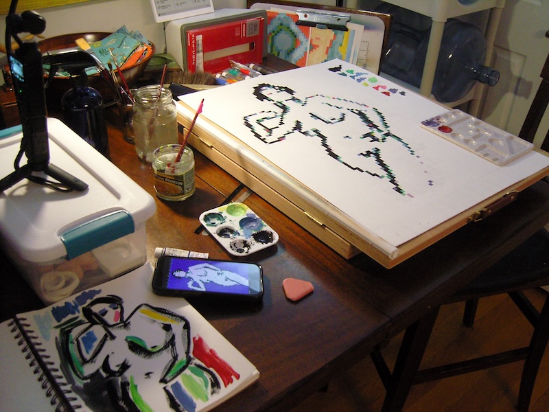 photo of the first pixel art study about halfway done, with the black outline pixels and the different collor accent pixels. you can also see my sketchbook where i'm using the excess paint to paint anotehr figure, my phone showing the digital reference, and a camera recording the process