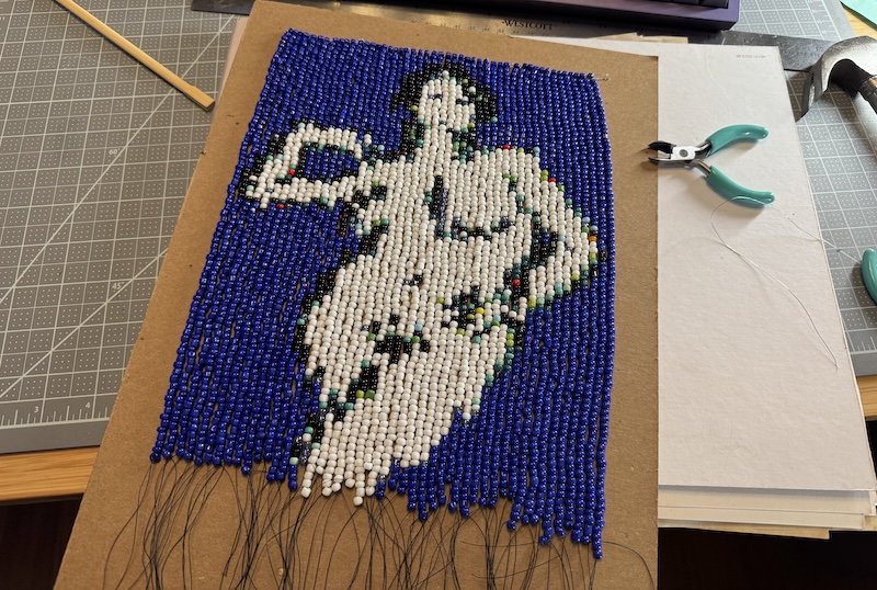 photo of a 48*80 size 6 seed bead tapestry mounted to a piece of brown binder board. it's all loose strands connected on the top row, and it measures around 8 by 11 inches