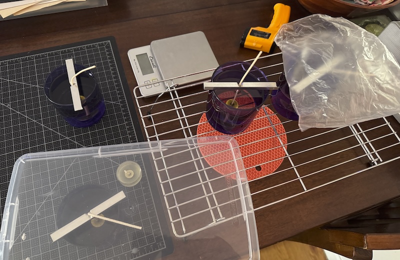 photo of a rack with some of my lavender smoke periwinkle glass candles curing. other tools like my scale and laser thermometer are on the table too