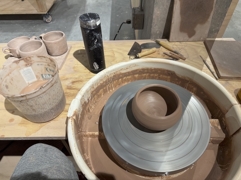 my station at the ceramics studio where i threw a nice bowl. in the background are my tools, my coffee thermos, a bucket of water and clay slip, and a board with three dry mugs i made a couple weeks ago