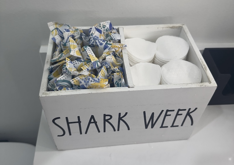 photo of a wooden bin on a sink that says "shark week" and on one side are tampons and the other side are cotton rounds