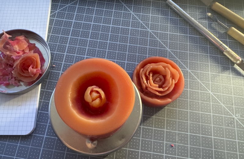two hand-carved flowers in beeswax, one of them is in progress. they are sitting on my gray desk mat alongside some carving tools and a tin lid holding beeswax pieces from my carving