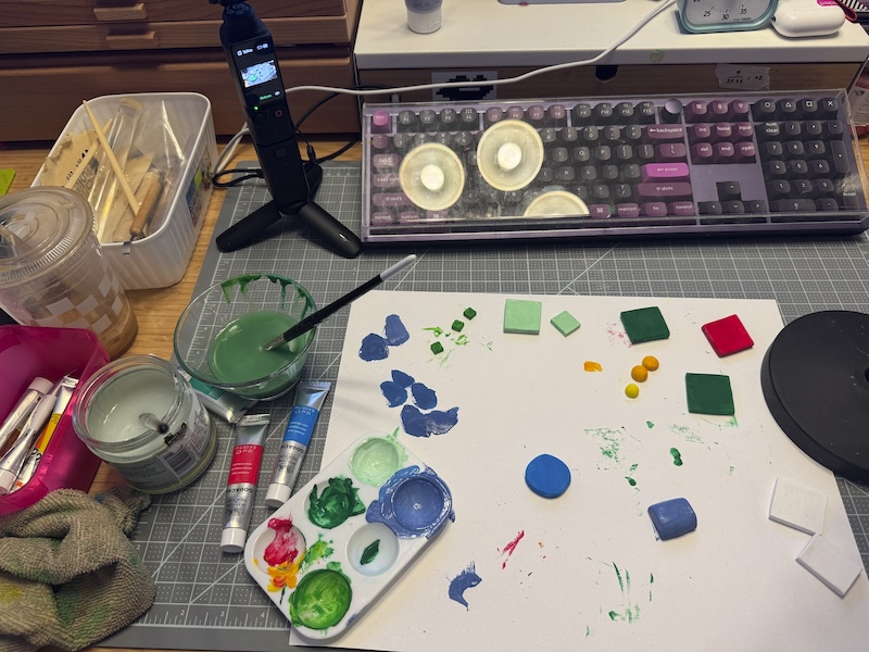 photo of my desk with my keyboard covered and a mat covered in air dry magnet pieces i was painting, alongside a palette of paint and my tubes and waters