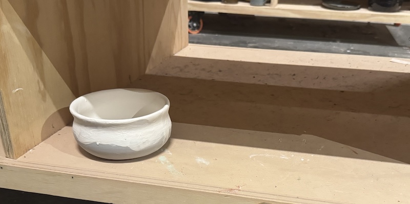 a photo of my glazed and yet to be fired mini cauldron bowl on the ceramics studio shelf