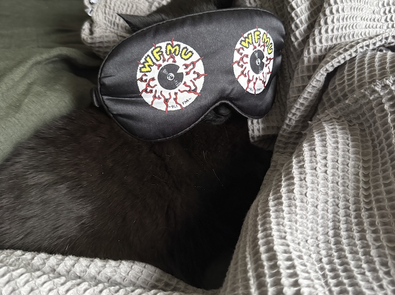 photo of my black cat pumagreg sleeping in a pile of my gray robe and green sheets. over his head is an eye mask that says wfmu and has two white vinyls with red veins to look like eyes