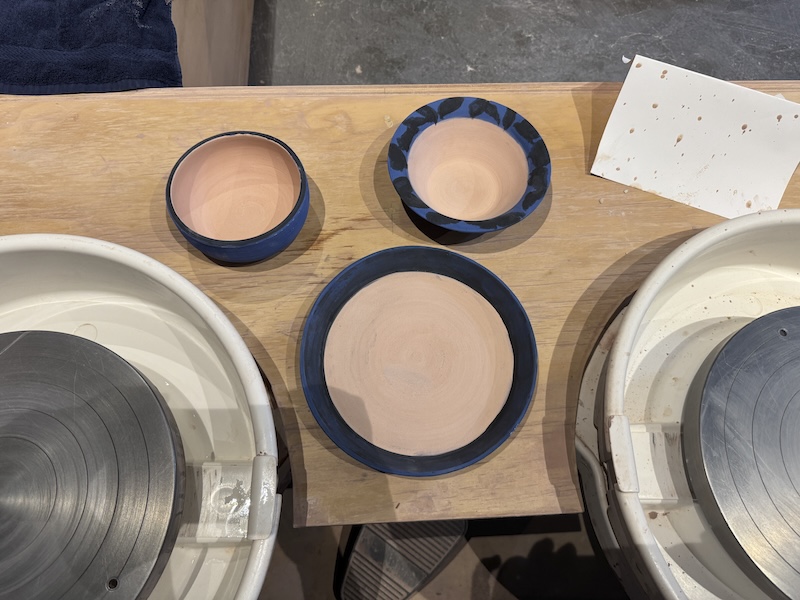 photo of three bisqueware pieces of mine next to my pottery wheel: two bowls and a blate with black and blue underglaze painting but the inside is blank still