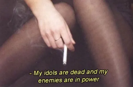 a photo of stockinged legs crossed with a hand holding a cigarrette captioned "- my idols are dead and my enemies are in power"
