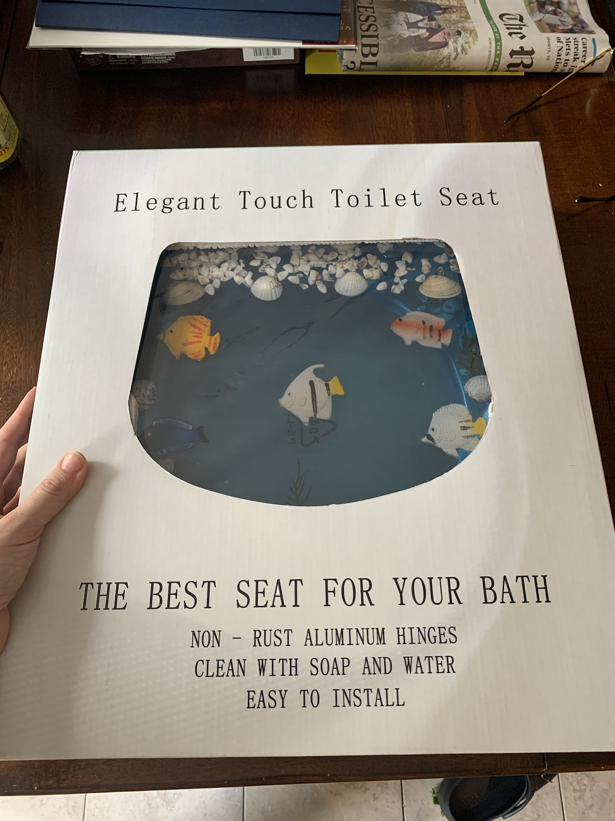 a photo of toilet-seat-sized box that says "Elegant Touch Toilet Seat" and there's a cut-out in the box exposing the toilet seat. Under the cut-out it says "THE BEST SEAT FOR YOUR BATH. NON-RUST ALUMINUM HINGES, CLEAN WITH SOAP AND WATER, EASY TO INSTALL"