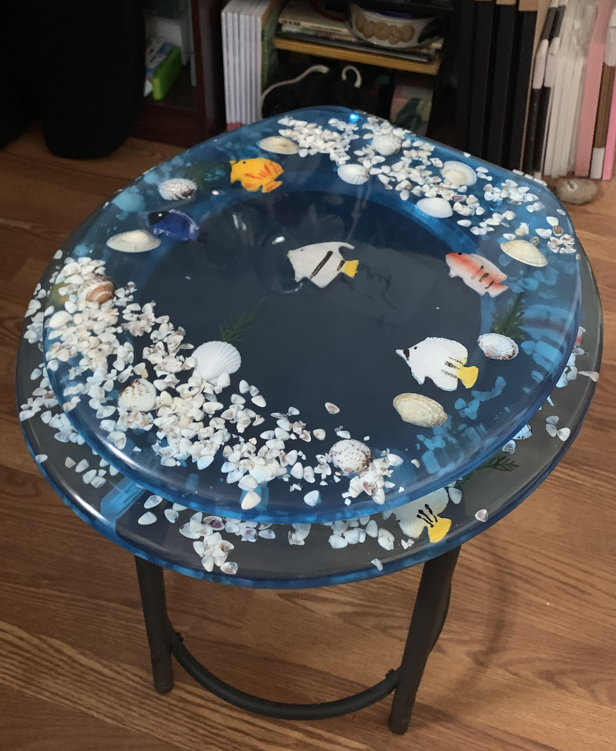 a photo of my new toilet seat, made of blue resin encapsulating fish and seashells, closed and on a black stool in my office