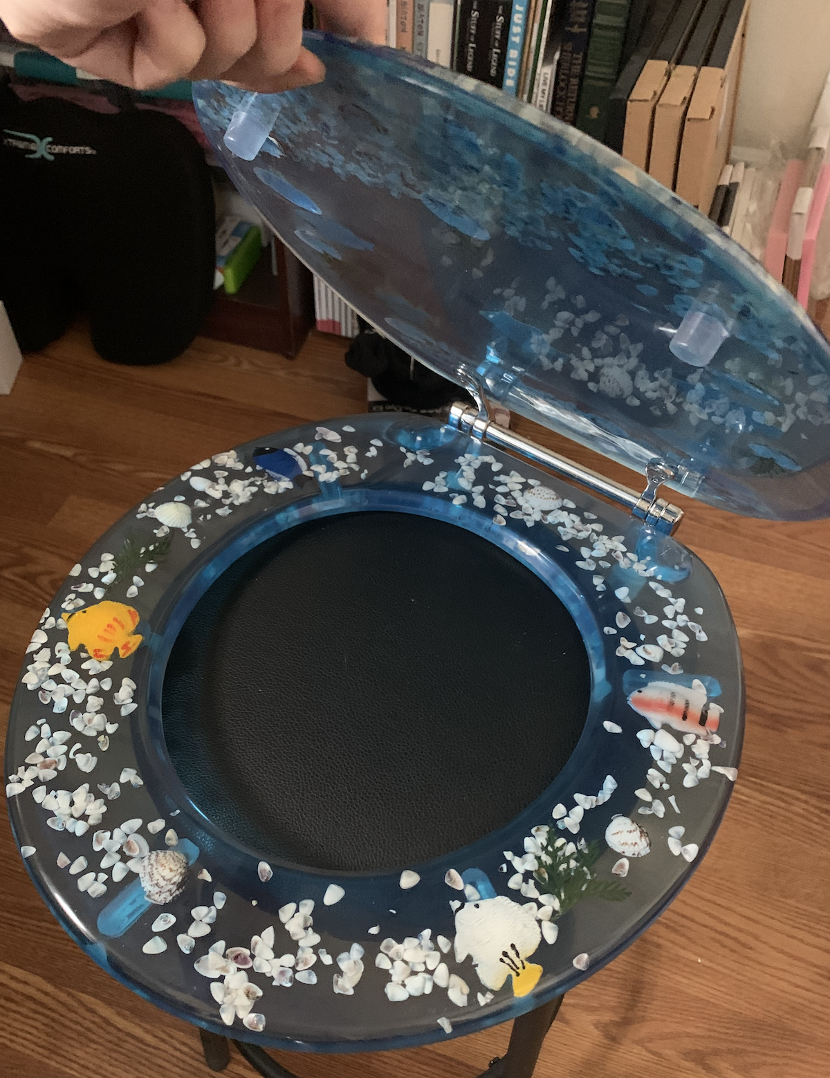 a photo of my new toilet seat, made of blue resin encapsulating fish and seashells, open on the black stool in my office 