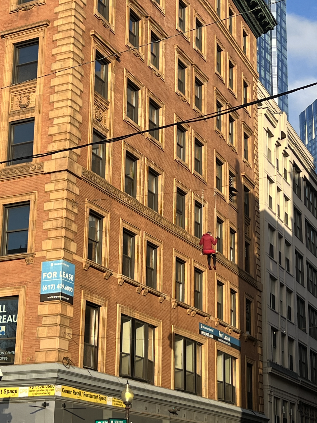 photo of between two buildings on boston common and there's a wire between the two and what looks like a woman on a swing a few stories up in the air but it's definitely not a real woman - but it looks like one!