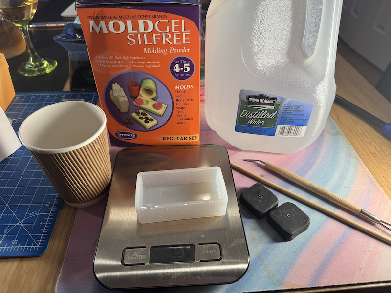 photo of my desk showing the materials i used to make the mold: moldgel box, scale, cup, clay scraping tools, the polymer version of what i'm molding