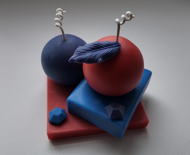 photo of a candle i made on a white backdrop. the candle has a red square base and a blue rectangle at an angle on top. there's a purple sphere on the back of the red base, and a red sphere with purple leaf on top of the blue rectangle. there's also a blue gem on the base and a purple gem on the blue rectangle