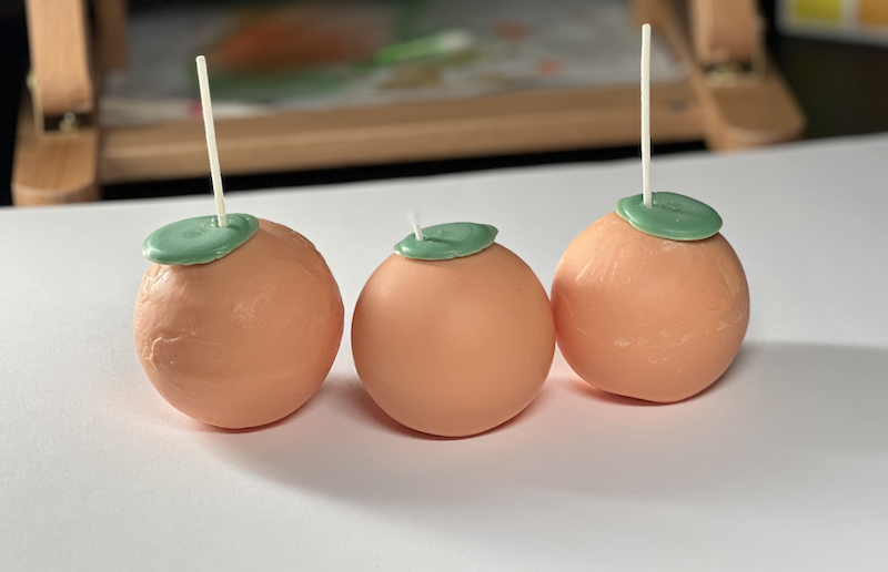 three orange fruit candles sitting with each other on a white background. 2 of them are more imperfect than the other, the wax is not as smooth