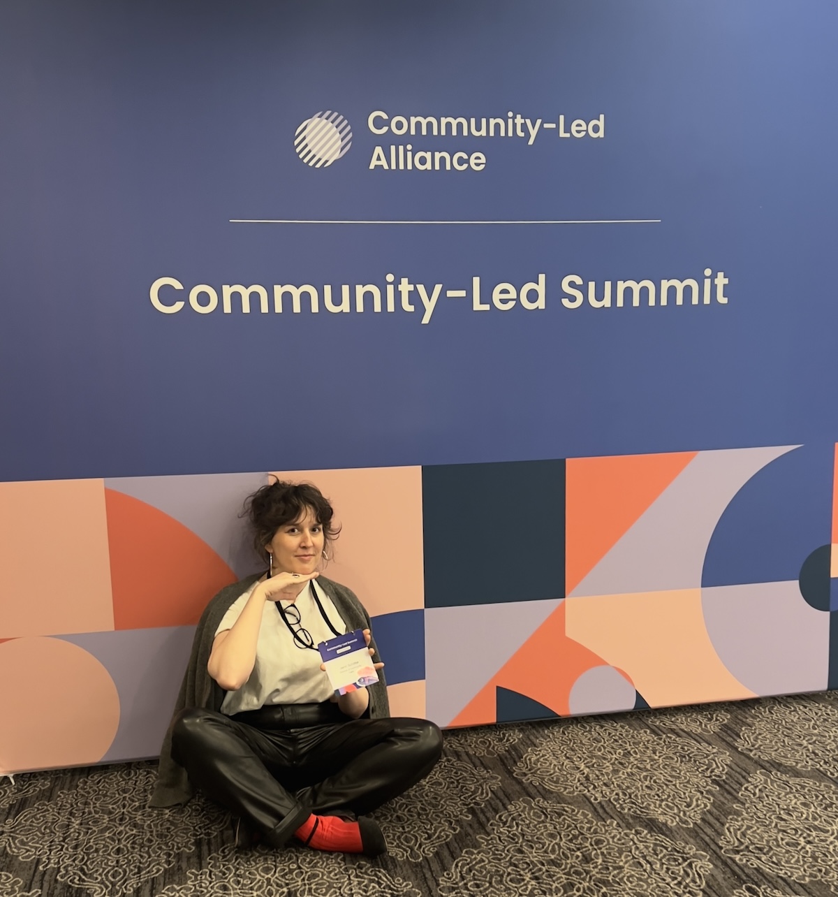 photo of me on the floor in front of a large banner that says "community-led alliance, community-led summit" in a drab conference room but i'm posing all cute, hand under chin like i'm a sweet angel