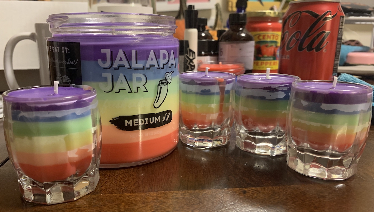 photograph of our final candles on my kitchen table, one is large and in a salsa jar and 4 are small shot glasses. they are rainbow wax going from red to purple in 6 layers