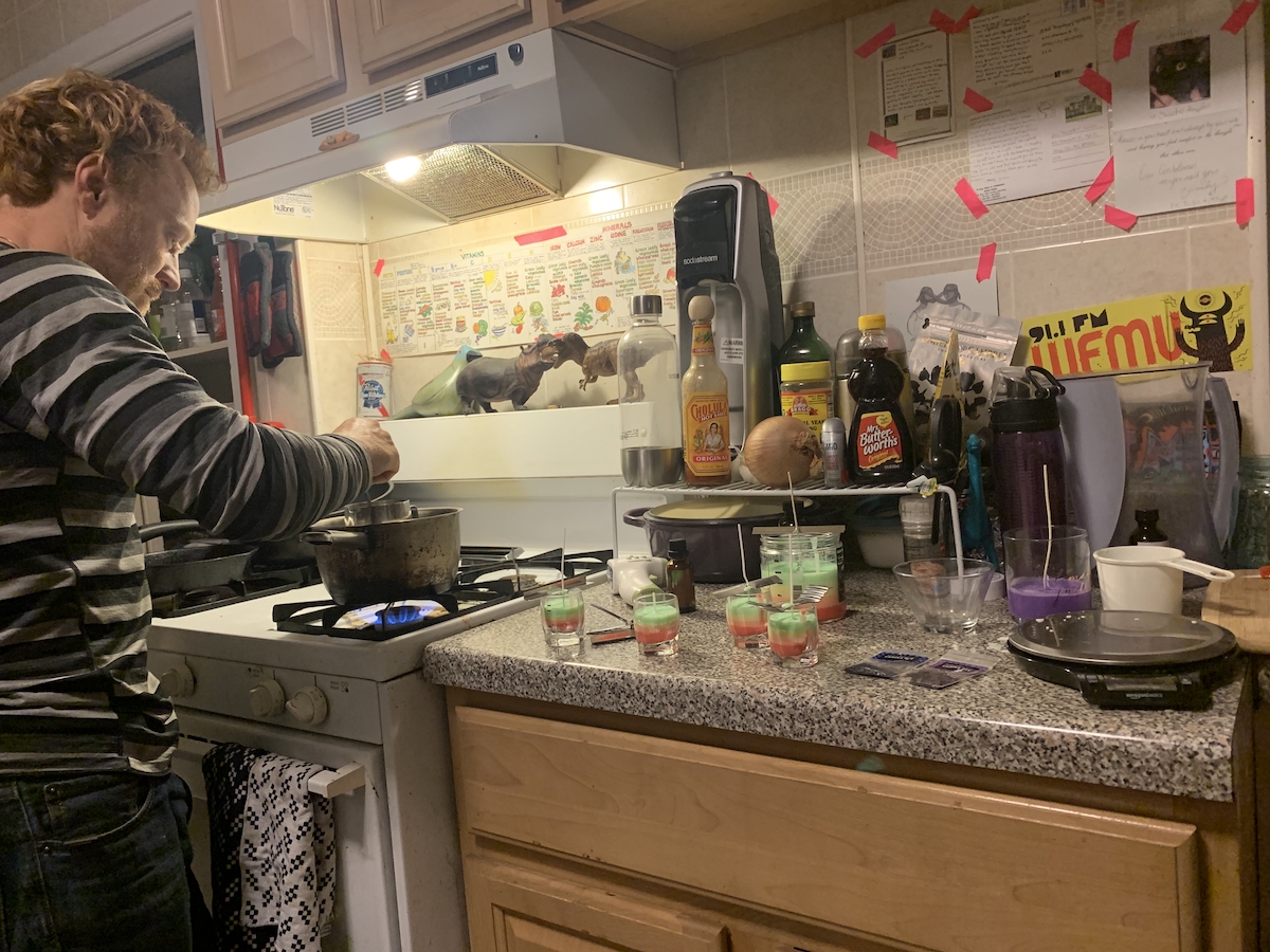 photograph of tyler stirring our double boiler of wax at my stove next to the counter where we're pouring our candles