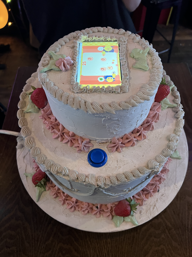 photo of a fake, pretty, two-tiered birthday cake and there's a mobile device screen built in on the top of it with a blue arcade button.